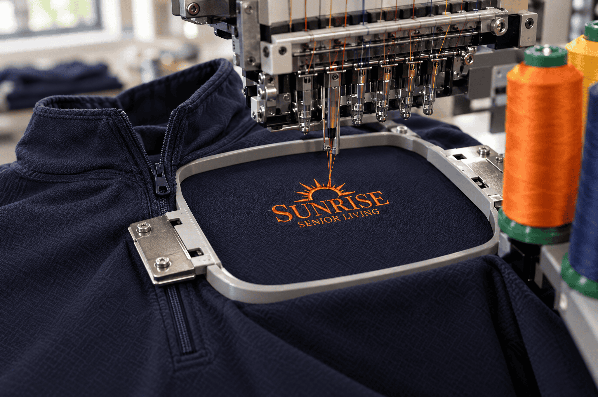 Embroidery machine stitching the Sunrise Senior Living logo onto a navy quarter-zip