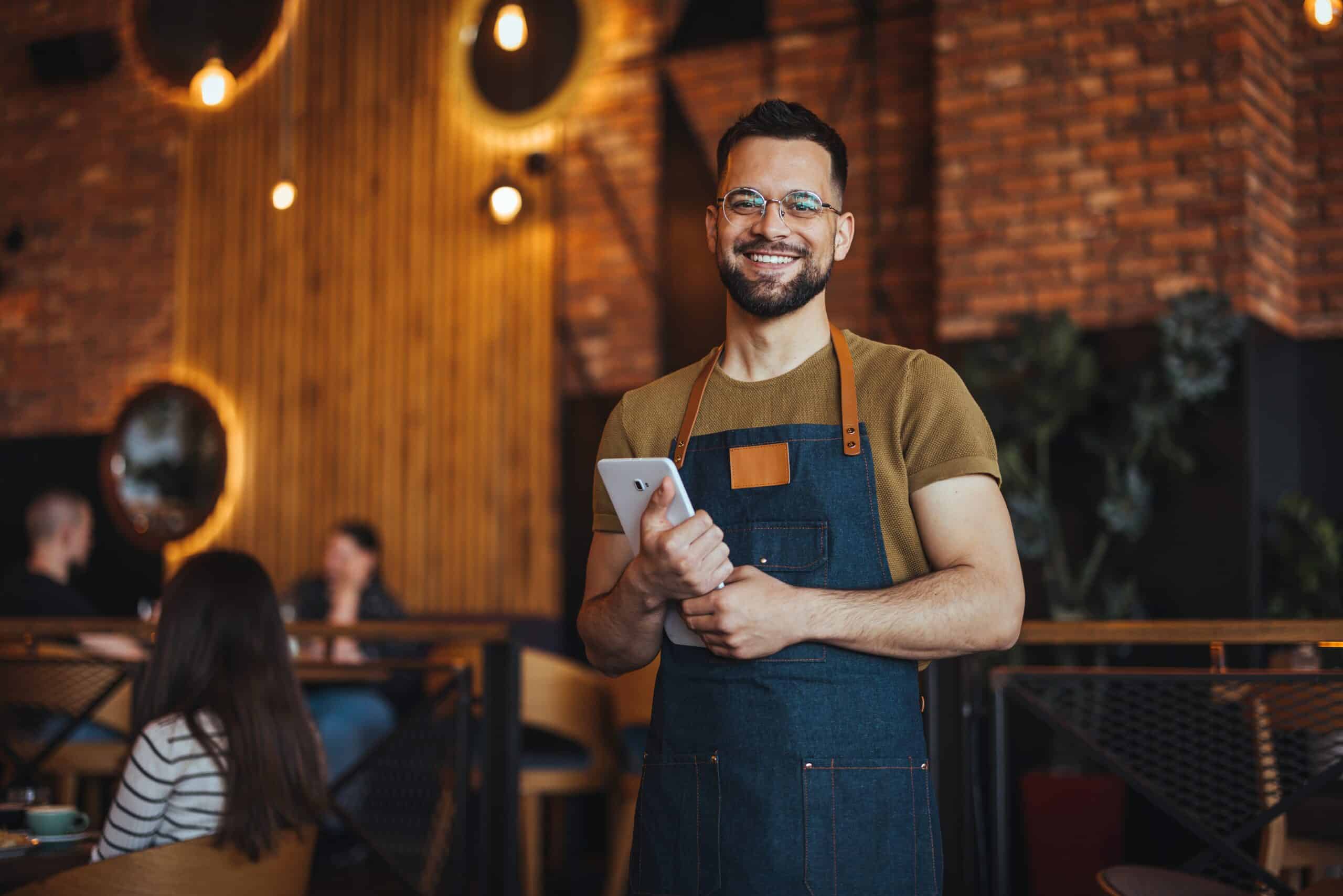 A smiling restaurant server in a denim apron holding a tablet